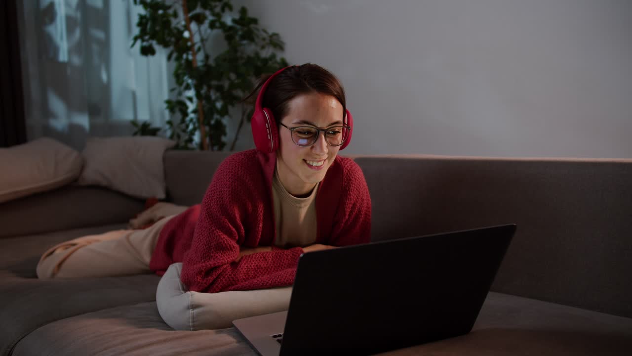 joven morena feliz con gafas en auriculares rojos inalámbricos y un suéter se acuesta en un sofá gris y estudia lenguas extranjeras con la ayuda de profesores en línea y lecciones en un apartamento moderno