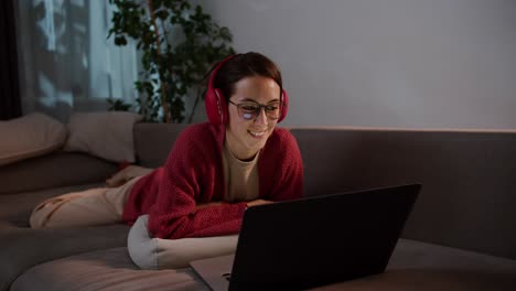 Happy-young-brunette-girl-with-glasses-in-red-wireless-headphones-and-a-sweater-lies-on-a-gray-sofa-and-studies-foreign-languages-with-the-help-of-online-teachers-and-lessons-in-a-modern-apartment