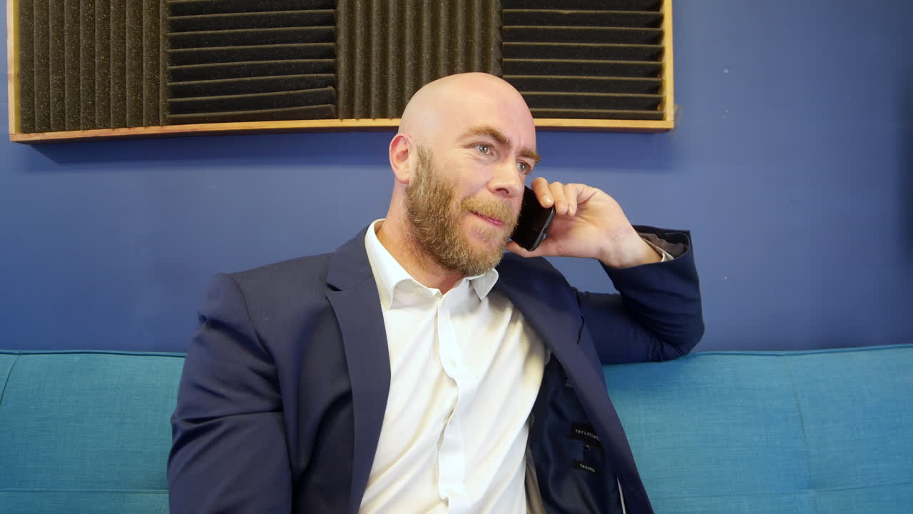Uomo d'affari calvo che ride e parla con un amico al telefono cellulare mentre è seduto sul divano nell'ufficio domestico