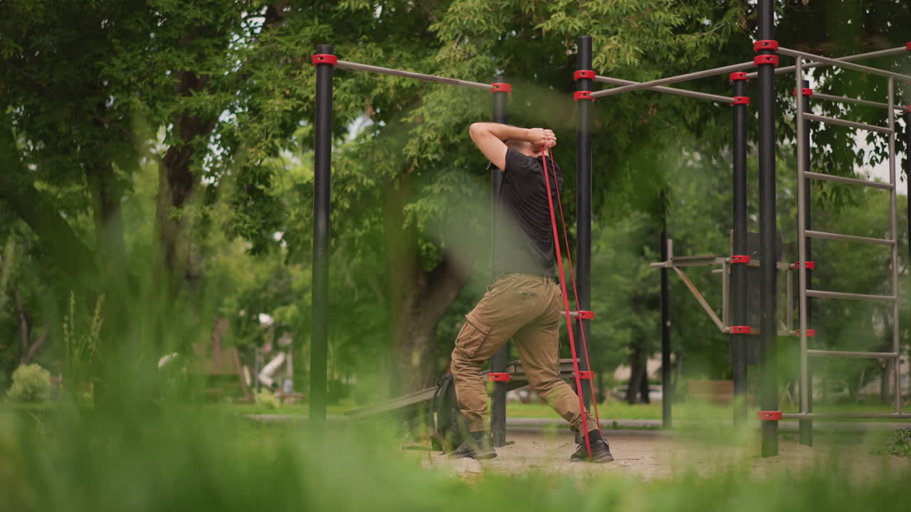 Man Exercises Outdoors, Individual Doing Resistance Band Assisted Pullup At Outdoor Fitness Station, Man Engaged In Stretching Exercise Using Resistance Band At Leafy Recreational Outdoor Gym