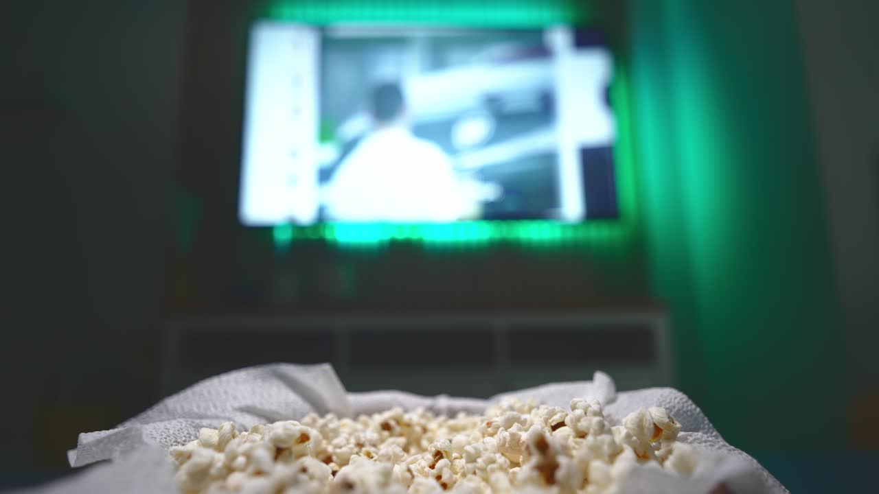 Lähikuva puukulhosta, joka on täynnä kotitekoista popcornia elokuvaillaksi. Käsi kurkottaa sisään ja ottaa popcornia, viihtyisä kotipurtava, rentoa viihdettä ja hauska perheen tai ystävien kokoontumishetki.