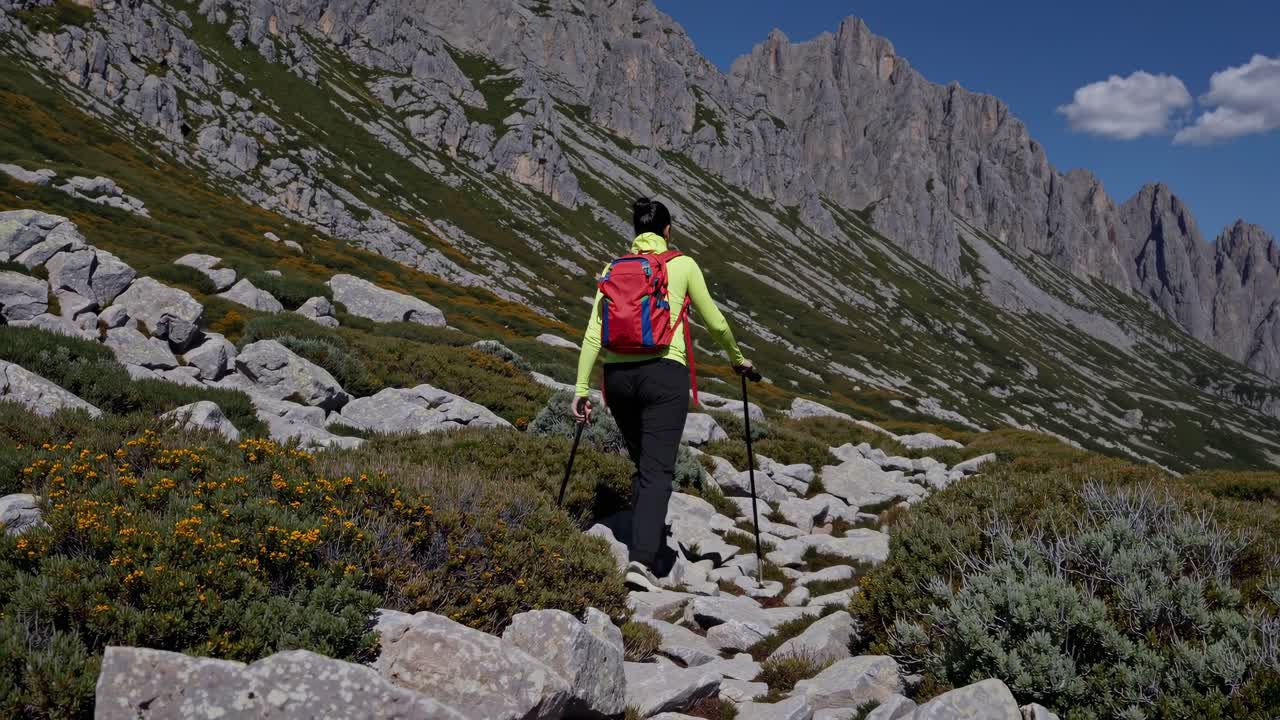 A hiker with a red backpack walks on a rocky mountain trail. Captured from a low-angle, the video