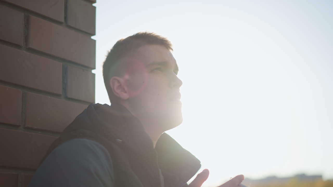 sunlight beams from behind man leaning on brick wall as he raises head with cigarette in hand, smoke curling around his face in bright glow creating dramatic silhouette atmosphere