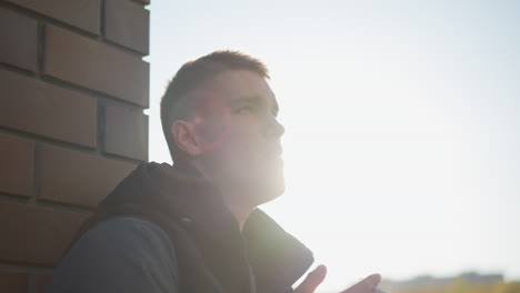 sunlight beams from behind man leaning on brick wall as he raises head with cigarette in hand, smoke curling around his face in bright glow creating dramatic silhouette atmosphere