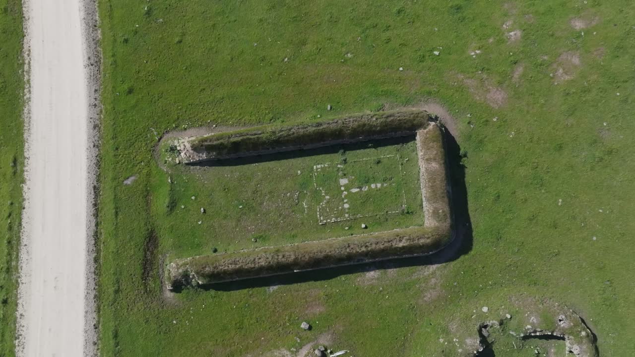 Drone descending in top-down view over 18th-century stone structure for pig farming. Feeding and watering trough remains visible inside. Surrounded by fields and dirt road.