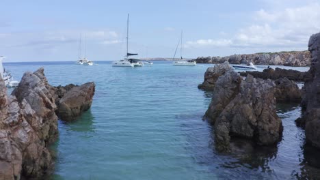 Catamarans-anchor-off-the-rocky-coastline-as-drone-flys-between-rocks-to-reveal-yachts
