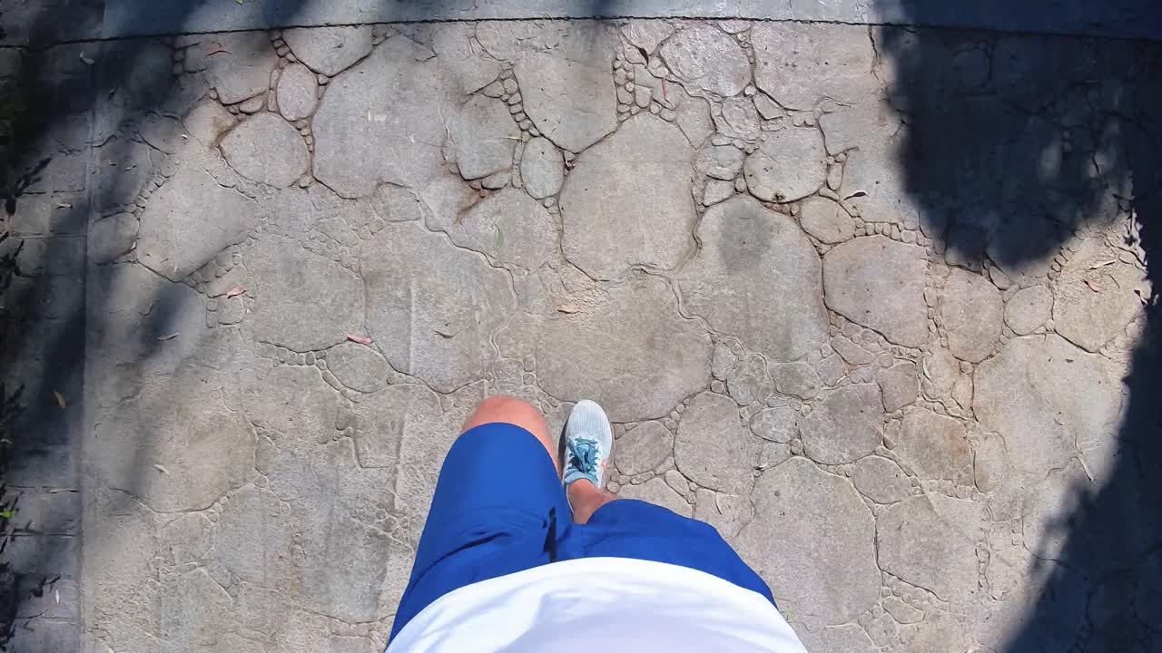 A top-down view of a person walking on a stone pavement in shorts and sneakers, casting a shadow in the sunlight. Ideal for themes related to travel, fitness, outdoor activities, and casual walking.