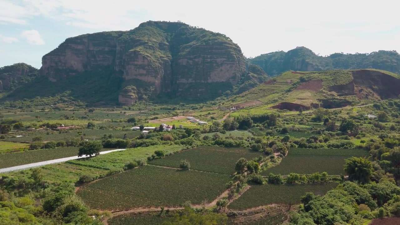Paesaggio rurale panoramico con terreni agricoli verdi e cime rocciose caratteristiche sopra campi coltivati e alberi sotto un cielo nuvoloso con un sentiero sterrato in primo piano e terreno aperto