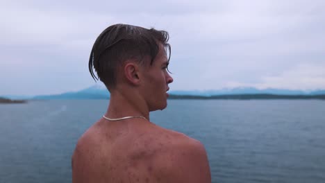 toma en cámara lenta de un joven con agua goteando de su rostro, usando un collar de plata, mirando el hermoso paisaje marino, tomando el momento y abrazando la sensación de vida y libertad