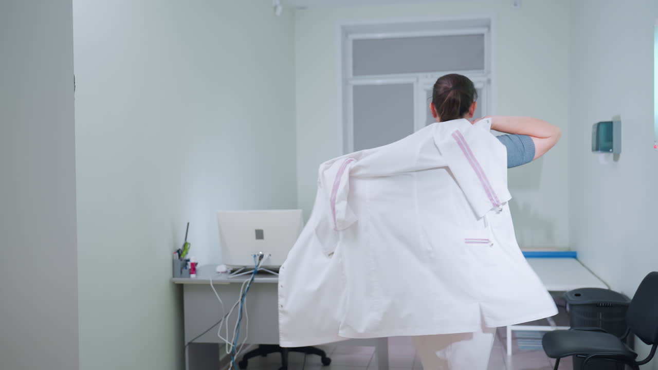 Back view of nurse wearing crocs putting on uniform while walking toward monitor in clean medical room equipped with desk chair and examination bed, ready to begin duties in well lit workspace