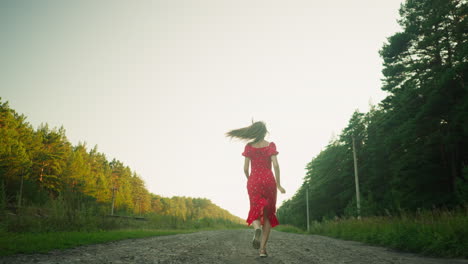 woman in red floral dress running on unkept dirt road through peaceful countryside, surrounded by green forest trees on both sides, backlit by soft natural light under clear sky during golden hour