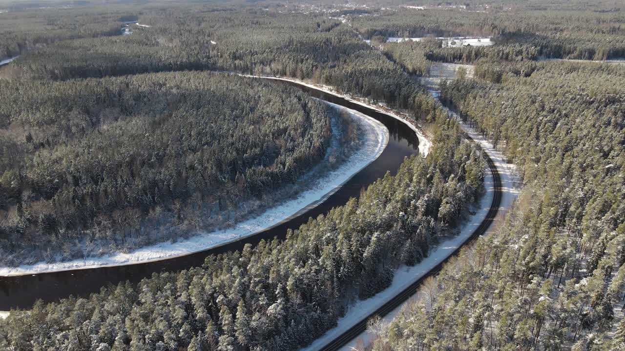 Aerial view of a winding river through a dense snow-covered forest in winter, with a curving road running alongside. A serene and scenic natural landscape.