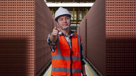 asian male engineer with safety helmet smiling and touching his chest then pointing at you while standing with red brick packed in stacks are stored