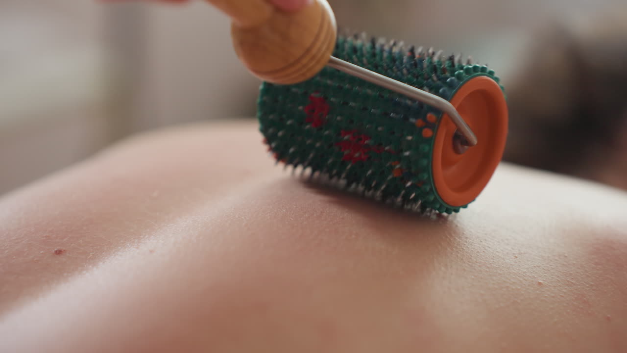 Close-up view of massage roller with wooden handle pressed on fair skin woman's back during relaxing therapy session, textured tool stimulating muscles with blurred soft light backgroumd
