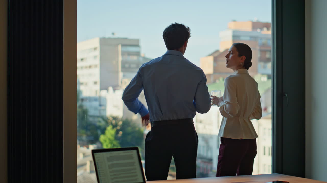 Office colleagues sharing coffee standing at panoramic window. Relaxed coworkers