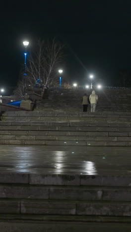 nighttime view of two people walking up a wide, illuminated urban staircase, with tranquil ambiance of the city at night, and street lamps with architectural details