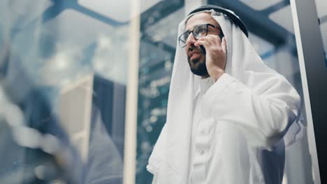 successful muslim businessman in traditional white kandura riding glass elevator to office in modern business center. man talking on a phone in a lift. saudi, emirati, arab businessman concept.