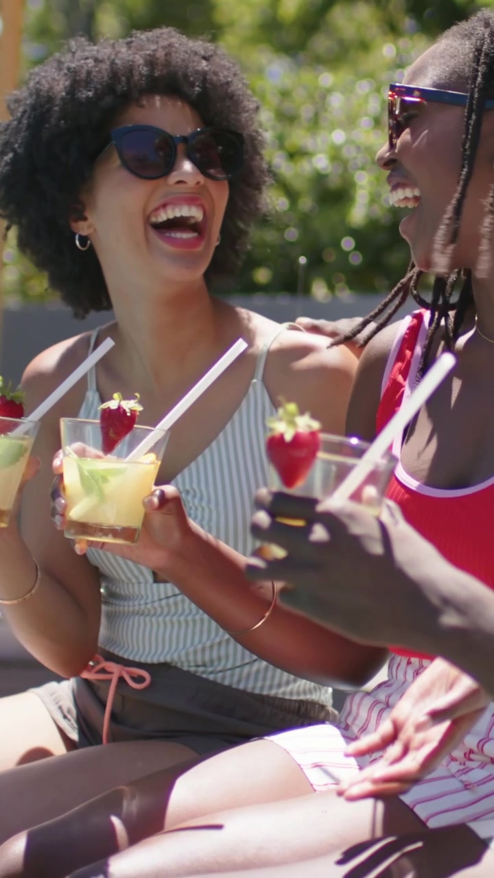 Vertical video: Camera framing woman in striped top raising drinks to relax with friend at poolside