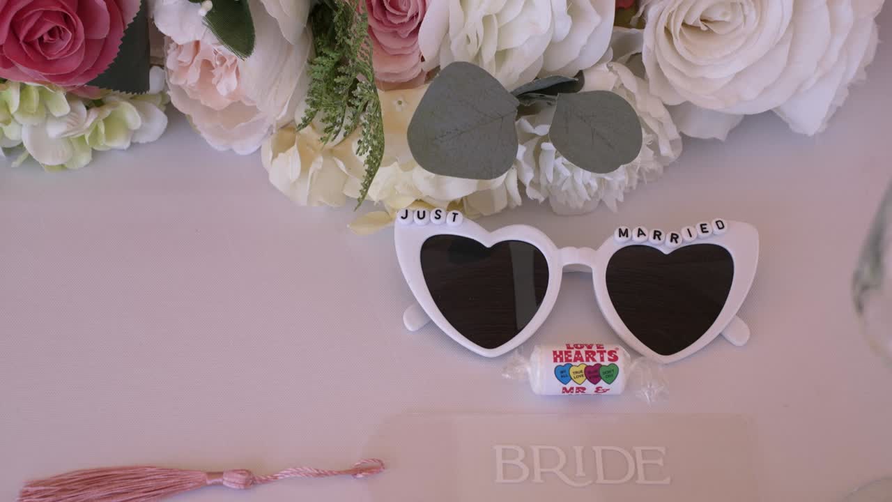 Heart-shaped sunglasses labeled just married rest on a table adorned with beautiful flowers. Surrounding the glasses are touches of wedding decor, enhancing the celebratory atmosphere
