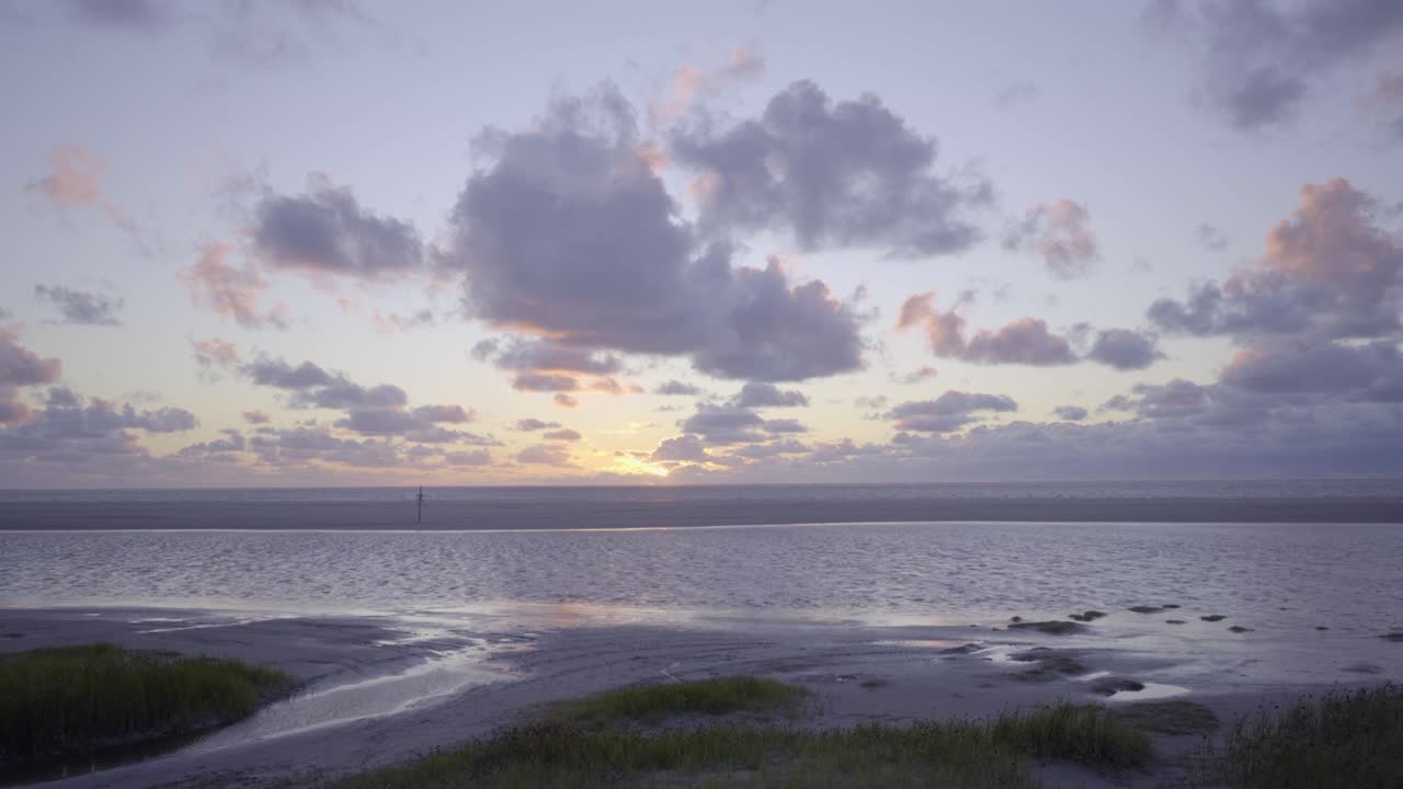 Forward and upward moving wide shot over long, green grass revealing the beach and ocean at Schiermonnikoog, the Netherlands. Sun setting against a blue, partly cloudy sky,