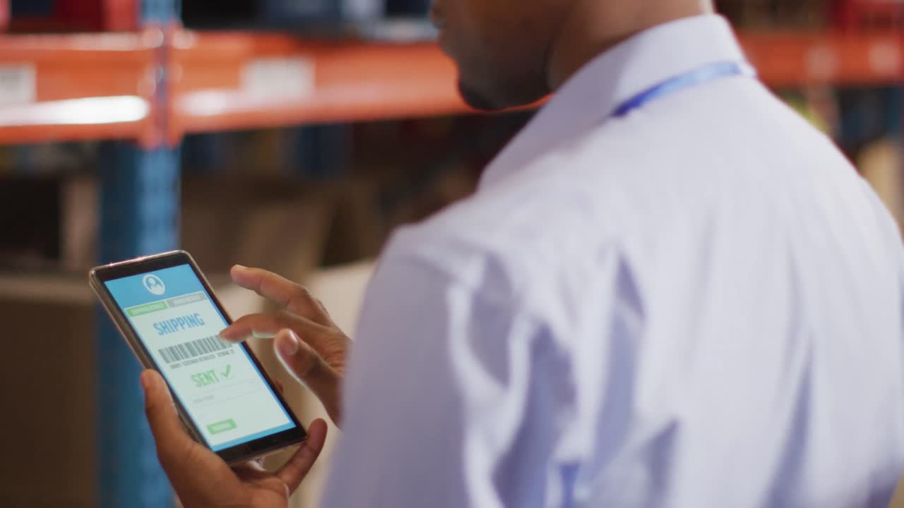 trabajador masculino afroamericano con casco usando un teléfono inteligente en un almacén