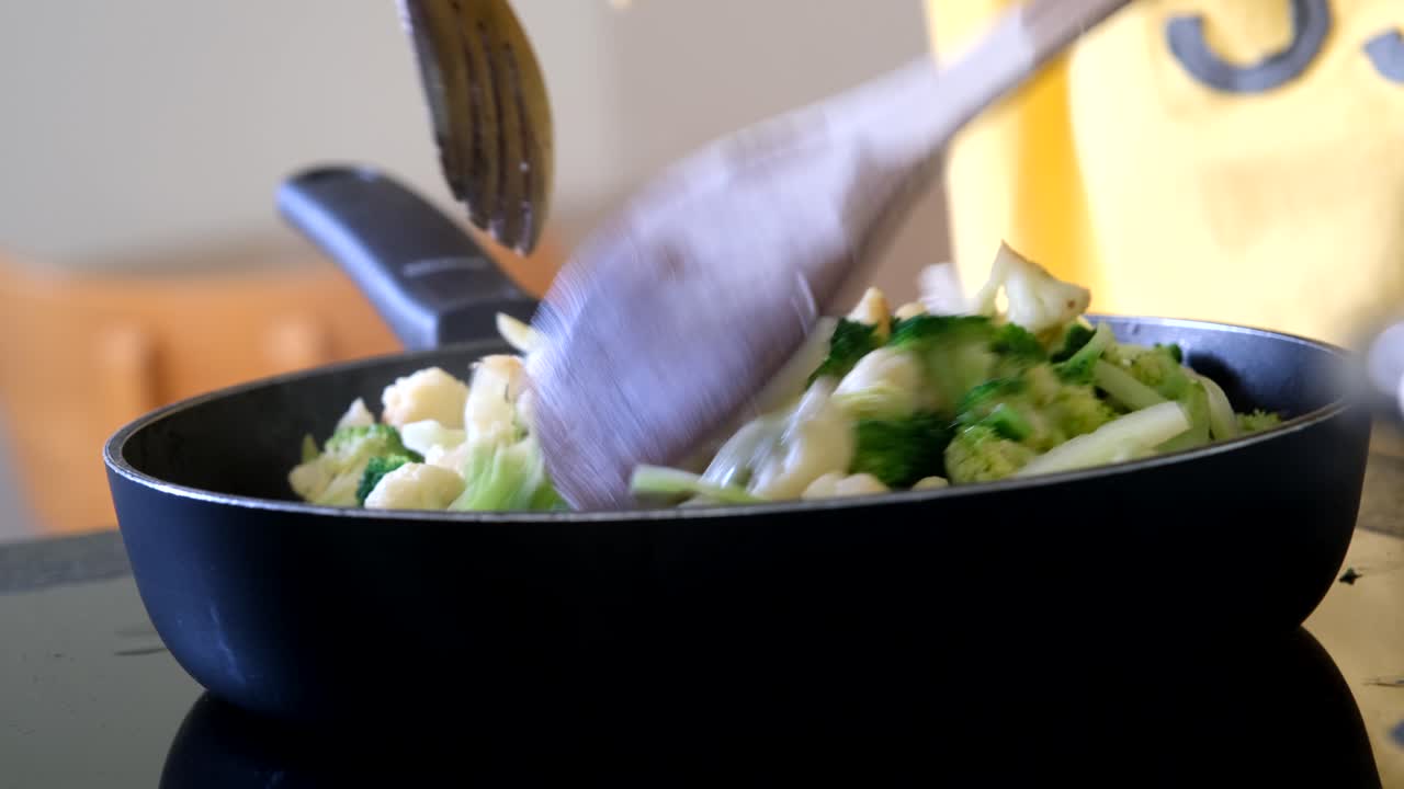 Fresh broccoli and cauliflower being sautéed in a black pan with a wooden spatula, capturing a healthy vegetable stir-fry in a home kitchen setting