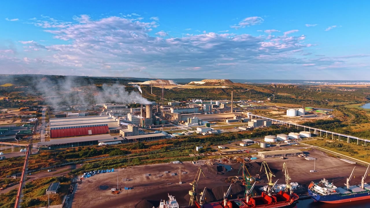 Kargo terminalinin yanında endüstriyel kompleks ve depolama tankları. Geniş panorama; fabrika binaları, konveyörler, liman vinçleri ve teraslı taş ocağı tepelerini içerir.