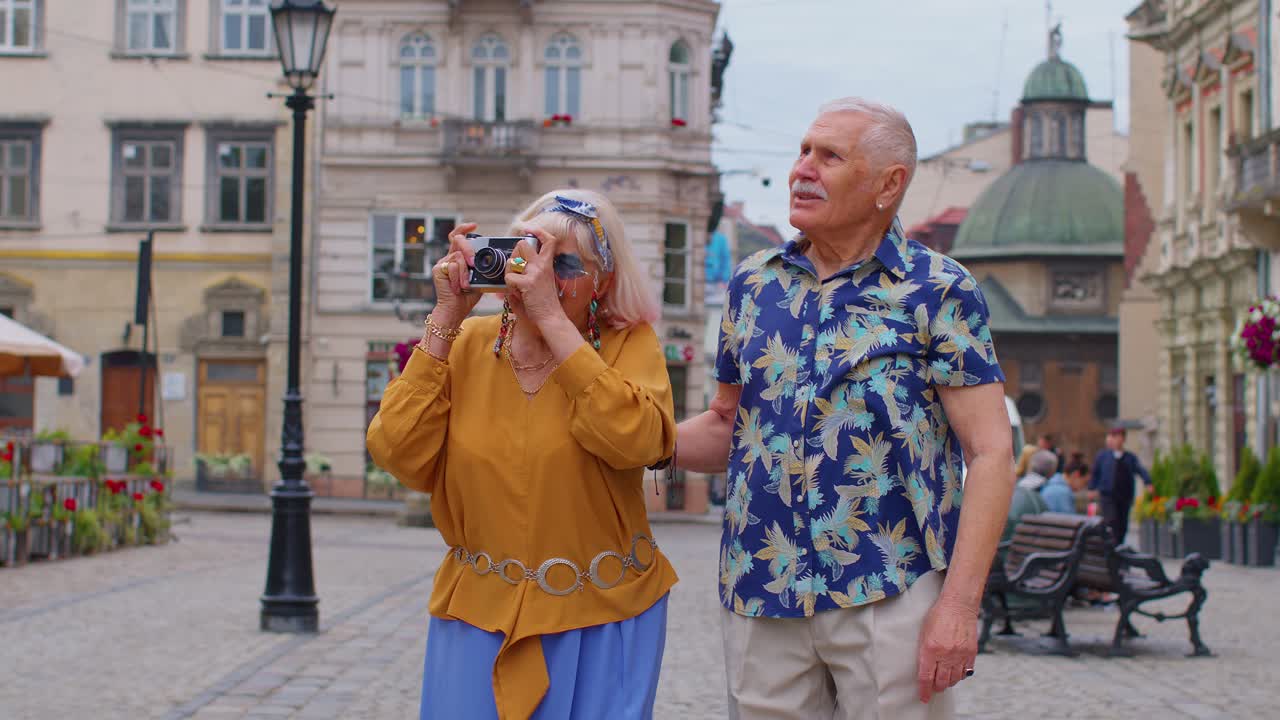 Senior old stylish tourists man woman walking, taking photos on old camera at summer city center