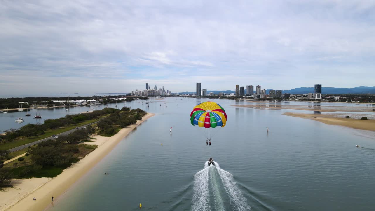 큰 다채로운 패러세일은 도시 스카이라인을 향한 해안 입구를 따라 스피드보트 뒤에 떠다니고 있습니다.