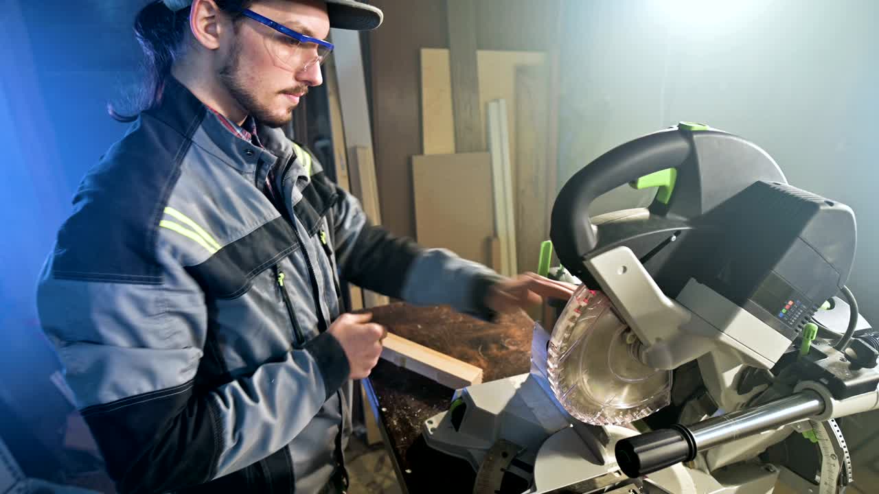 joven carpintero barbudo concentrado con gafas de protección y una gorra comprueba la pieza de trabajo