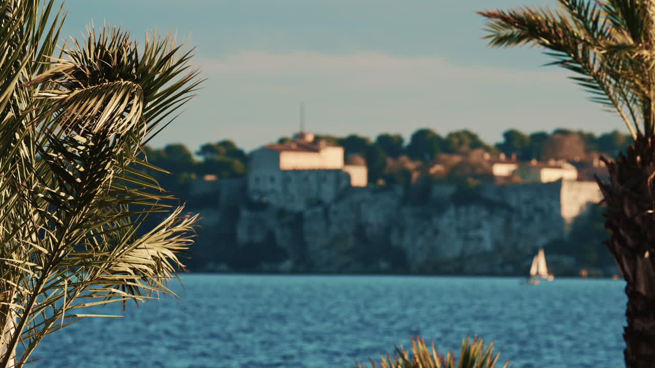 Palmetrær som svaier forsiktig i forgrunnen, med det historiske Fort Carre med utsikt over Middelhavet i Antibes