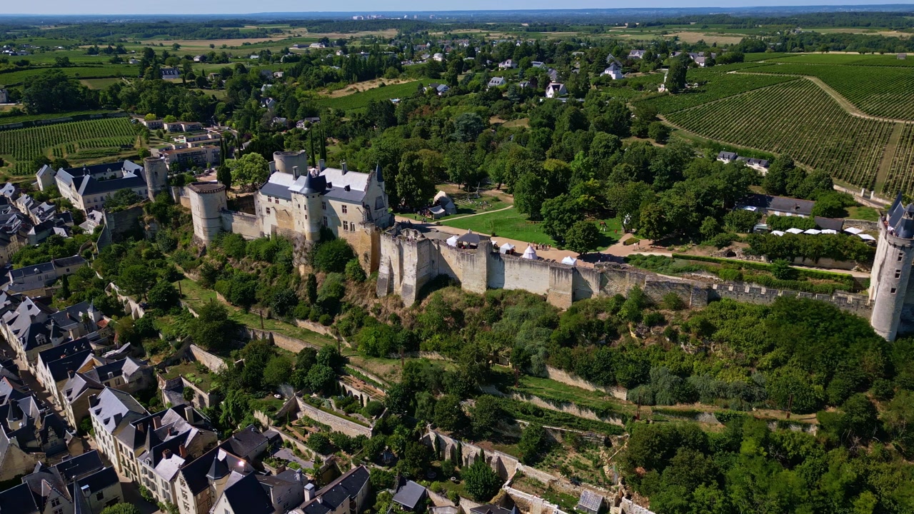 Lateral drone shot of Chinon’s royal fortress, showing stone walls, surrounding trees, nearby town, and open fields.