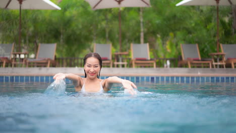 Una-Mujer-Joven-Y-Bonita-Que-Se-Relaja-En-La-Piscina-De-Un-Resort-Sonríe-Mientras-Salpica-Juguetonamente-El-Agua-En-El-Aire