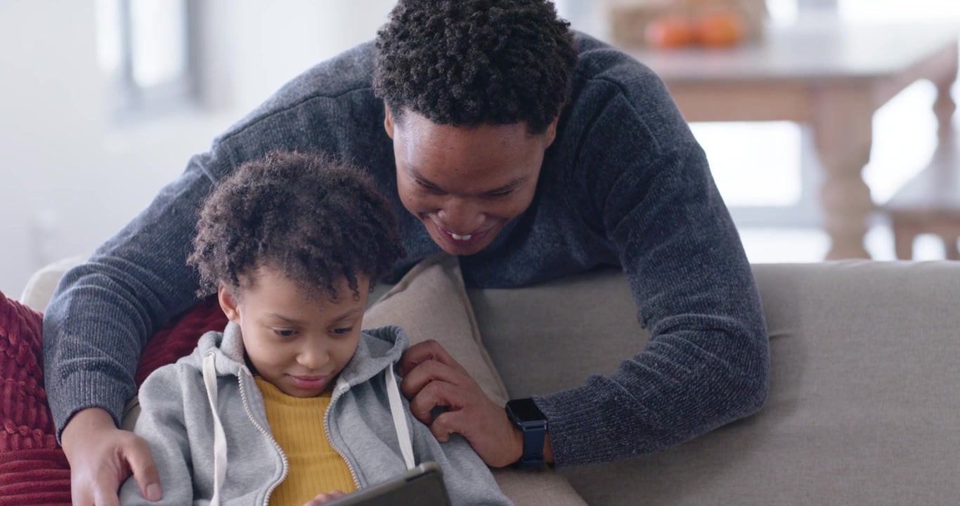 African American father approaching couch, steadying tablet, helping youth girl tapping screen