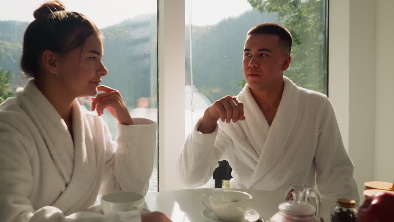 Guy throws rose petal into tea brewing in cup and winks to lady. Young couple in terry bathrobes has fun drinking hot beverage at breakfast together. Relaxed morning routine