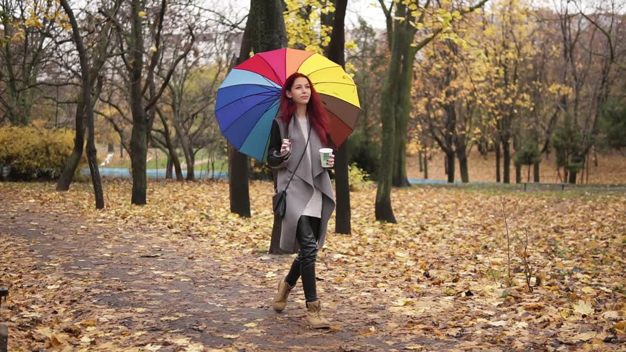 joven mujer elegante con el cabello rojo caminando en el parque de otoño y bebiendo café de una taza de papel mientras sostiene un paraguas de colores. niña con abrigo cálido disfrutando del clima de otoño fresco con una taza de bebida caliente