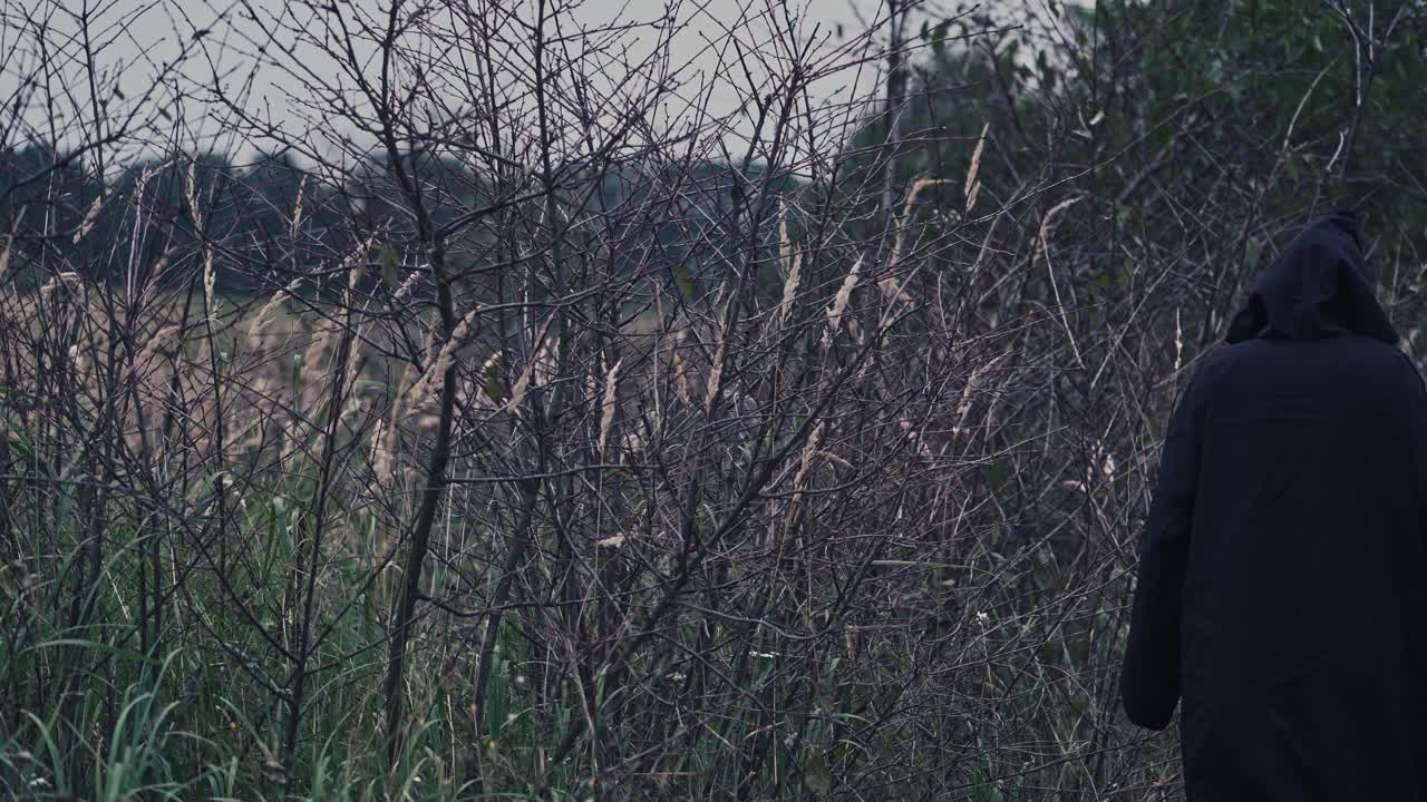 Back view of a dark figure outdoors. Ghost in black cloak going away on nature evening background.