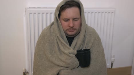 freezing young man wrapped in a blanket sitting next to a heater, shivering from cold and drinking tea