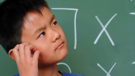 front view of thoughtful asian schoolboy scratching his head against chalkboard in classroom 4k