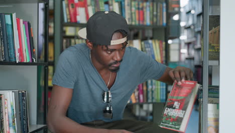 College student in casual tee with backward cap and sunglasses on shirt pulls book from shelf while seated between library rows, examining cover under soft indoor light surrounded by colorful spines