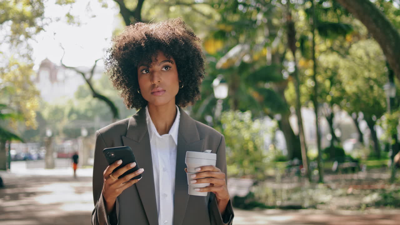Forretningskvinde kigger på smartphone, stående i bypark med papirkaffekop.