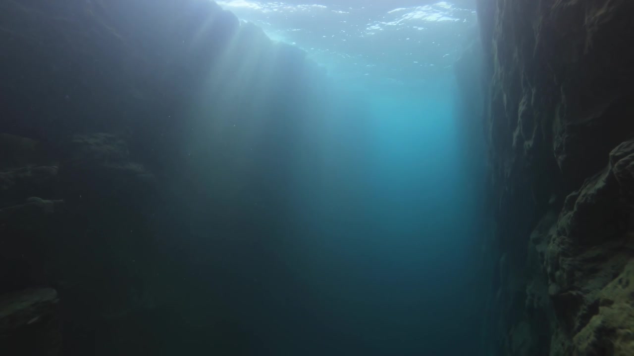 Coluna cintilante iluminada pelo sol entre paredes de rocha em cânion submarino, ondulações movendo feixes de luz e silte à deriva.