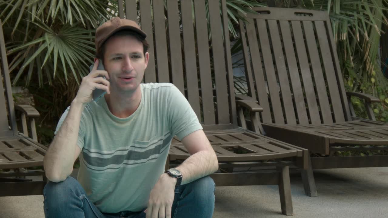 A man talks on the phone showing a range of emotion near beach chairs and tropical plants