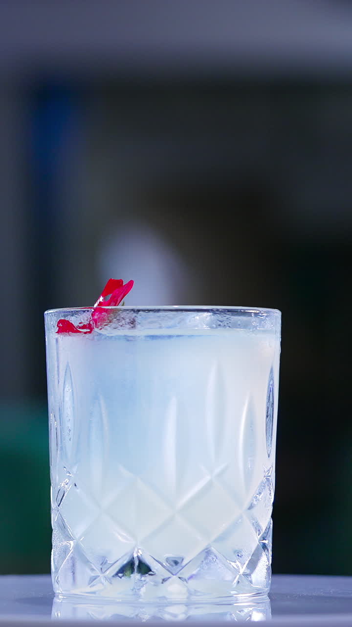 Old-fashioned glass with drink on the rotating table. Cocktail decorated with red flower in the bar. Close up. Blurred backdrop. Vertical video