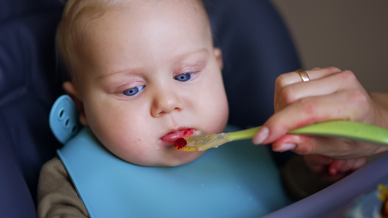 Mom's hand is holding a spoon in front of the baby face. Mother is feeding her child with fruit puree.