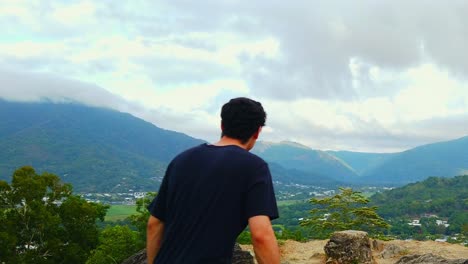 man reaches a lookout and stops to enjoy the view of the valley below in slow motion, a lush green valley and cloud covered mountains stretch out before him