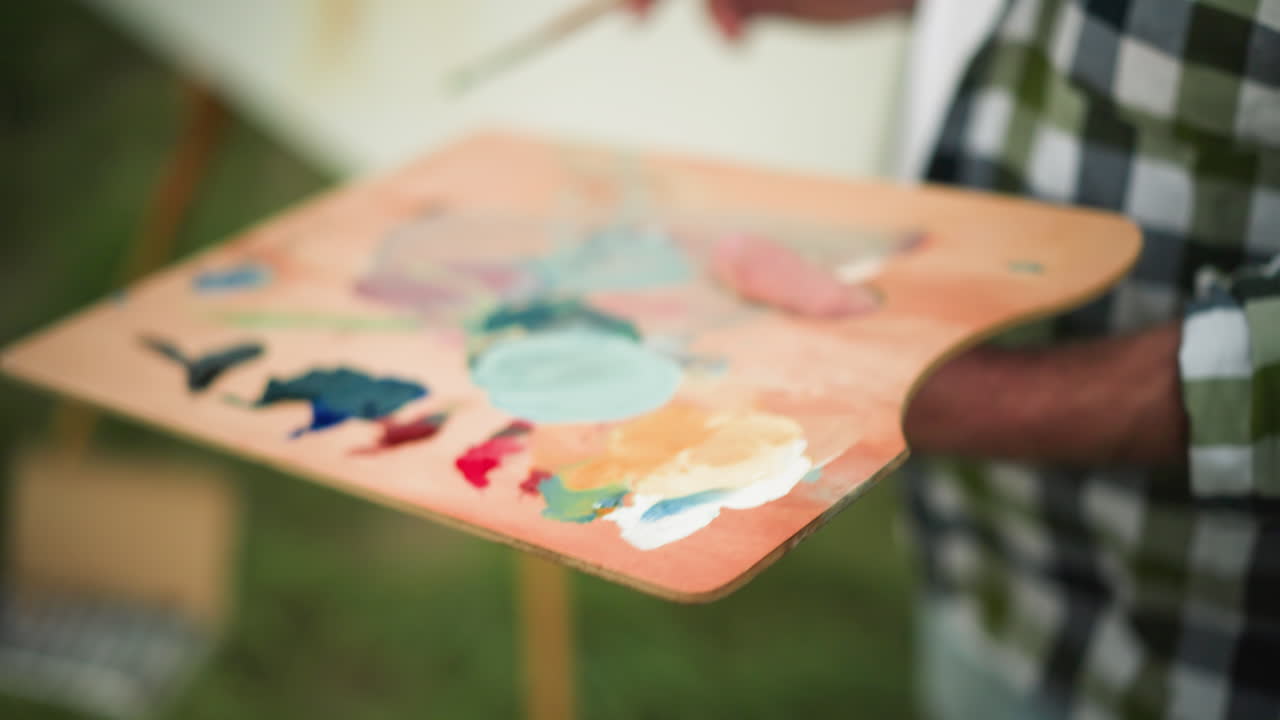 체크색 셔츠를 입고 얼굴이 보이지 않는 예술가 한 명이 화려한 색으로 가득 찬 페인트 브러쉬와 패을 들고 있습니다. 흐릿한 배경은  캔버스를 드러니다.