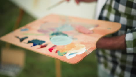 an artist wearing a checkered shirt, with their face not visible, holds a paintbrush and a palette filled with vibrant colors. the blurred background reveals a white canvas