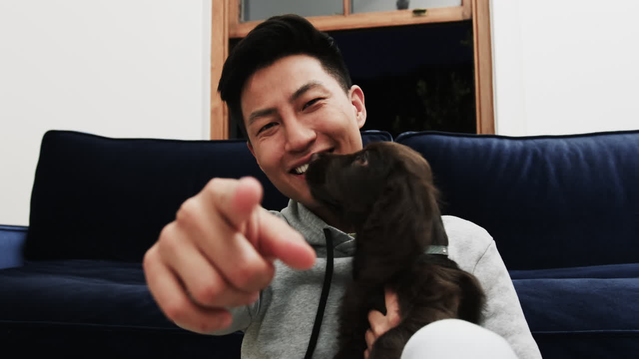 On video call, Smiling Asian man at home pointing while holding adorable dog on couch