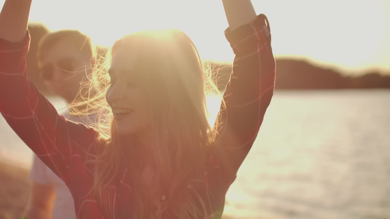 una estudiante está bailando en la fiesta al aire libre con sus amigas en la playa. su largo cabello rubio está volando en el viento. ella sonríe y disfruta de un verano en la costa del río con cerveza.